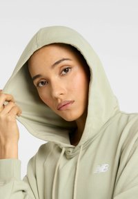Sweat �à capuche vert clair avec cordon de serrage, texture douce et logo brodé sur la poitrine gauche, porté tout en tenant la capuche.