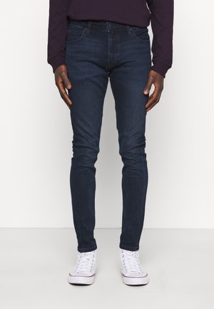 Homme portant un jean slim bleu foncé et des baskets montantes blanches, debout devant un fond clair uni.