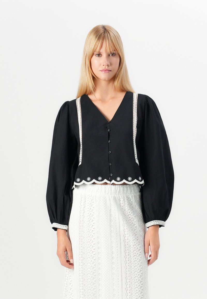 Blouse noire cropped avec des manches volumineuses, un décolleté en V, une fermeture à boutons à l'avant, et une bordure en dentelle blanche le long du décolleté et de l'ourlet, avec des bords festonnés.