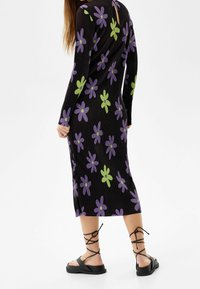 Czarna sukienka z prążkowanego materiału w purpurowe i zielone wzory kwiatowe, z długimi rękawami i wycięciem z tyłu. Założona z czarnymi sandałami na paski.