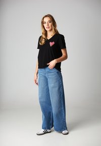 Czarna koszulka polo z dekoltem w serek z różowym sercem na lewej piersi; zestawiona z szerokimi niebieskimi dżinsami i białymi sneakersami.