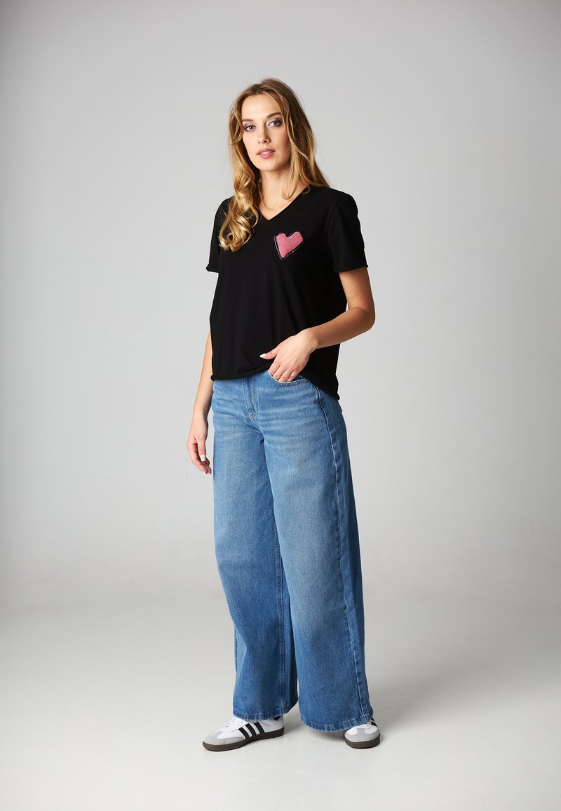 Czarna koszulka polo z dekoltem w serek z różowym sercem na lewej piersi; zestawiona z szerokimi niebieskimi dżinsami i białymi sneakersami.