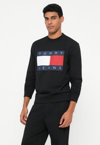 Hombre vistiendo un sudadera negra con un gran logotipo rectangular de Tommy Jeans en blanco, rojo y azul marino en la parte frontal, posando frente a un fondo blanco.