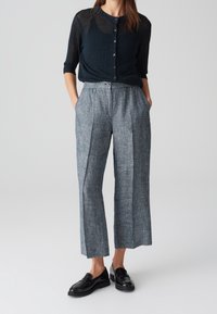 Femme portant un haut noir transparent à boutons, un pantalon large taille haute gris et des mocassins noirs, se tenant les mains dans les poches.