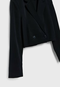 Blazer court noir avec larges revers et fermeture à double bouton, suspendu sur un fond gris uni.