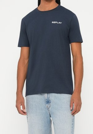 Homme portant un t-shirt bleu foncé uni à manches courtes avec un texte blanc "REPLAY" sur la poitrine et un jean bleu clair sur fond neutre.