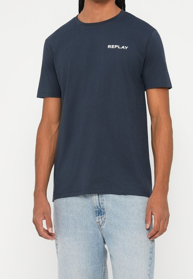 Homme portant un t-shirt bleu foncé uni à manches courtes avec un texte blanc "REPLAY" sur la poitrine et un jean bleu clair sur fond neutre.
