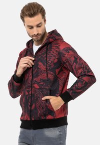 Rot- und schwarzfarbener Reißverschluss-Hoodie mit grafischen Drucken, ausgestattet mit einer Kapuze mit Kordelzug, Fronttaschen sowie gerippten Bündchen und Saum. Glatte Textur.