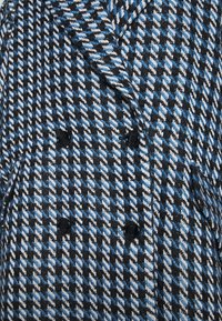 Dvouřadový kabát s černobílým a modrým houndstooth vzorem, se strukturovanou látkou a černými knoflíky.