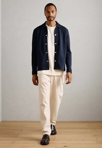Man walking indoors wearing a navy buttoned shirt jacket, cream t-shirt, cream pants, and dark brown loafers on light wooden floor.