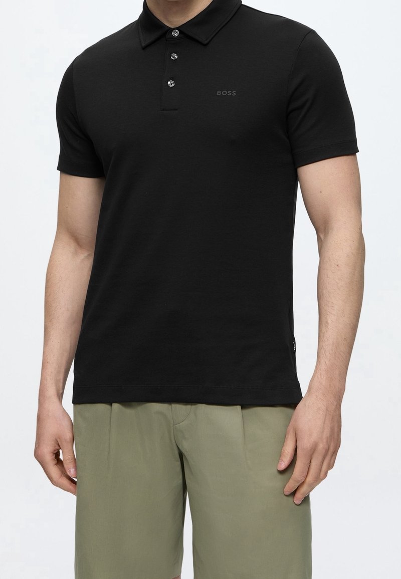 Homme portant un polo noir ajusté à manches courtes avec des boutons et un short vert olive sur un fond uni.