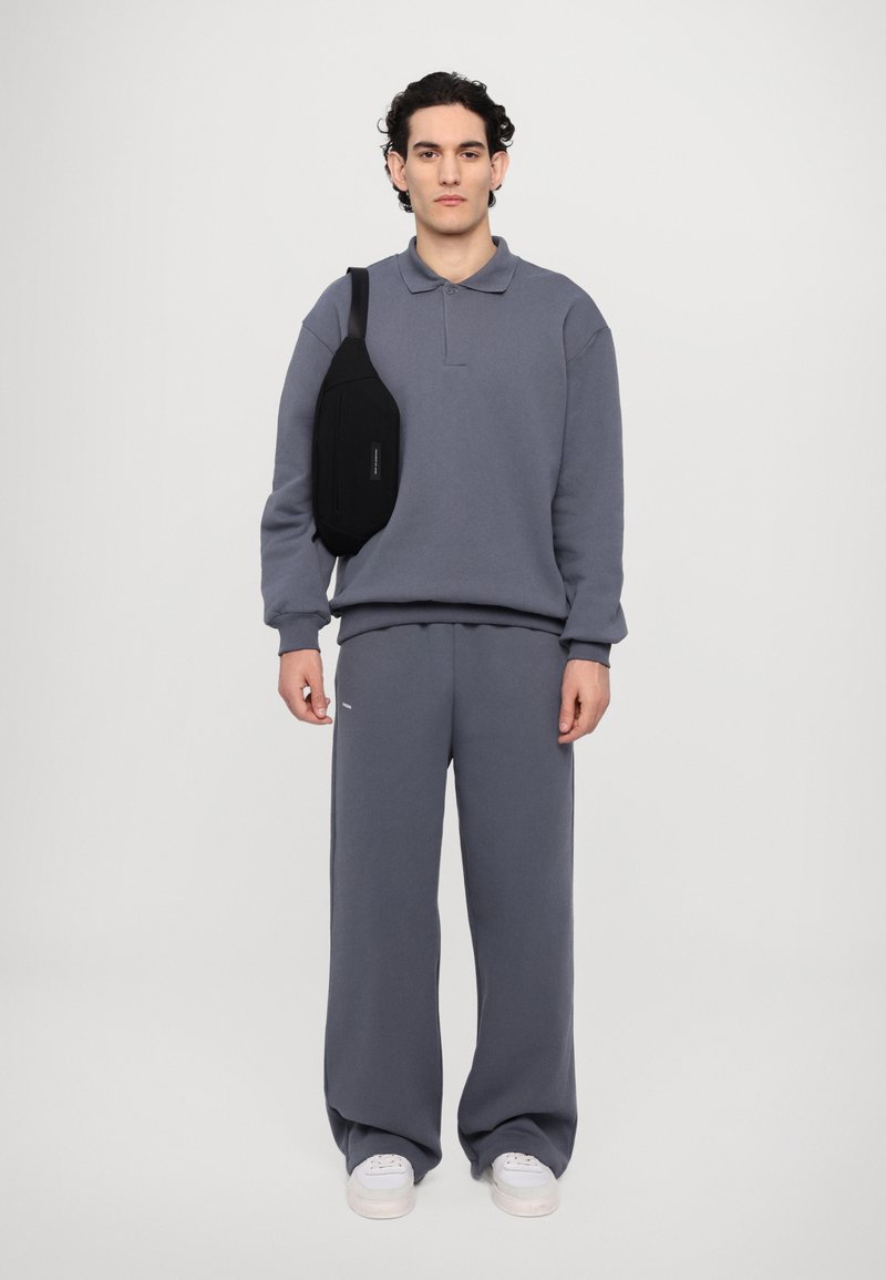 Šedá fleecová mikina s límečkem a odpovídající šedé tepláky. Černá taška nošená přes rameno, bílé detaily na teniskách.