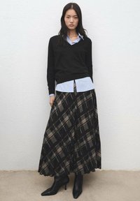 Čierny sveter s výstrihom do V cez svetlomodrú pruhovanú košeľu, doplnený dlhou kockovanou sukňou v čiernej, hnedej a béžovej farbe a čiernymi čižmami na opätku.