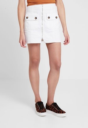 Frau trägt weißen Minirock mit Reißverschluss vorne und mit Knöpfen versehene Taschen, kombiniert mit Leopardenmuster-Sneakers vor weißem Hintergrund.