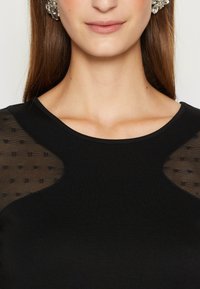 Frau in einem schwarzen Oberteil mit durchsichtigen, gemusterten Einsätzen nahe den Schultern und blumigen Ohrringen, sichtbar sind der Hals und der untere Teil des Gesichts.
