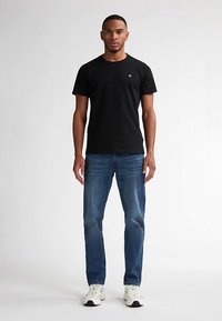 Svart bomulls-t-shirt med rund hals och liten logotyp på bröstet, kombinerad med blå jeans och vita sportskor.