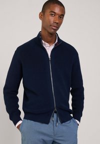 Granatowy sweter rozpinany z elastycznym splotem, srebrny zamek błyskawiczny, wysoka stójka i prosty dół. Noszony na białej koszuli w czerwone paski.