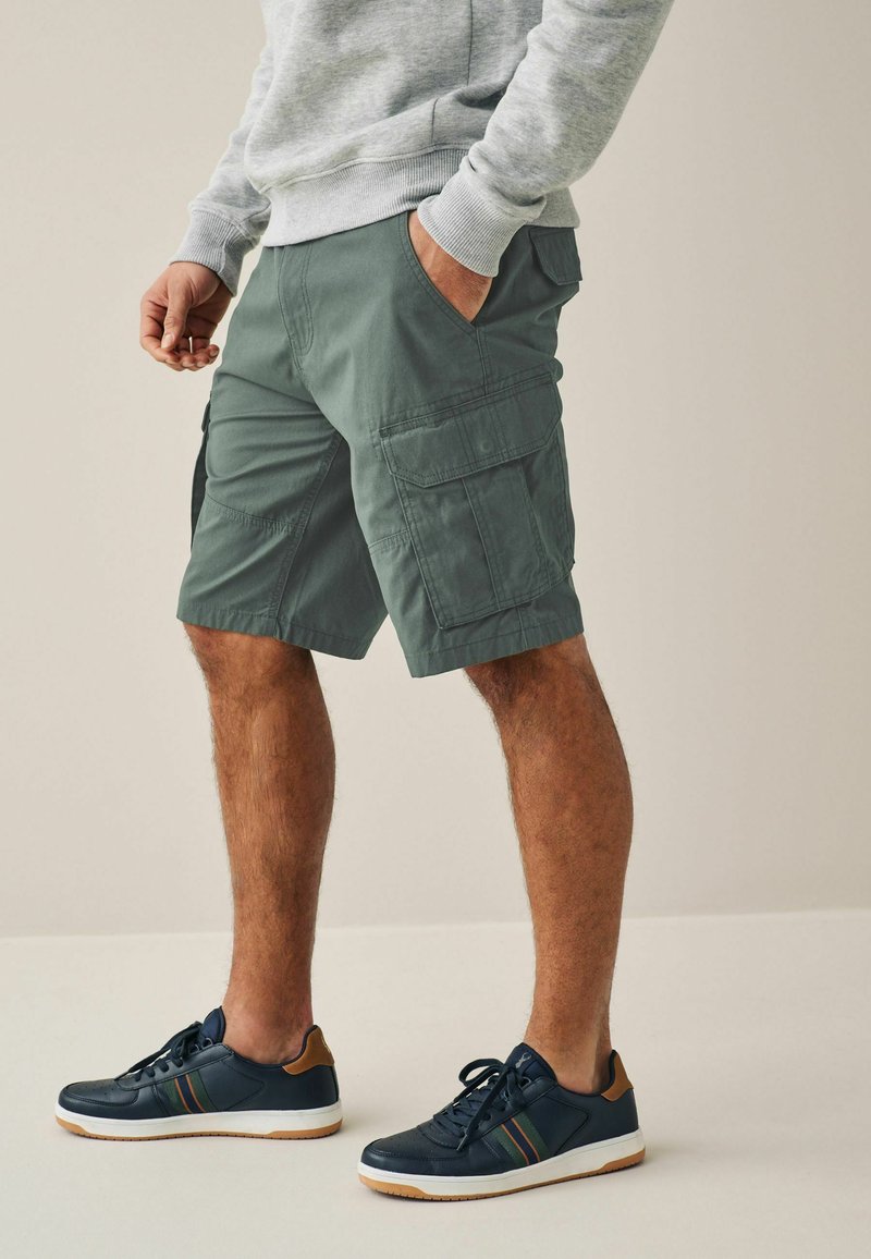 Shorts cargo en coton vert muet avec deux poches latérales et deux poches arrière. Associés à des baskets sombres avec un accent et des rayures marron.