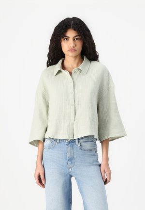 Lysegrønn cropped skjorte med knapper laget av teksturert stoff, med krage og vide ermer, kombinert med lyseblå jeans.