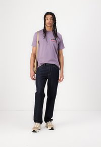 T-shirt en coton violet avec un logo de montagne, jeans en denim foncé et baskets beiges. Cheveux longs et tressés et sac en bandoulière beige.