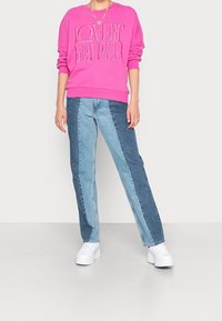 Růžová bavlněná mikina s nápisem "LOVERS REBEL", spárovaná s modrými džíny s kontrastními světlými a tmavými panely. Bílé platformové tenisky.