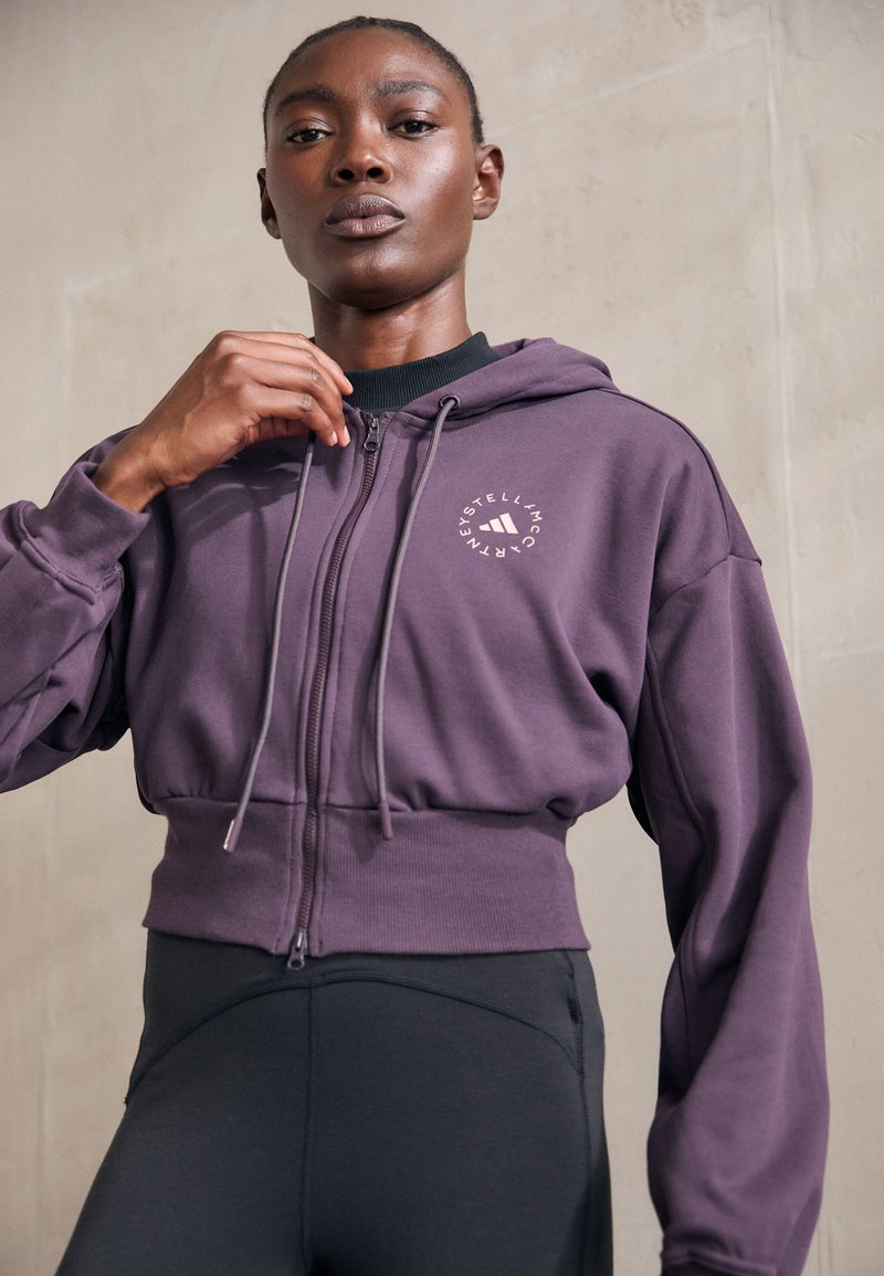 Cropped purple hoodie with a front zipper, drawstrings, and logo. Paired with black leggings, showcasing a smooth texture and athletic fit.