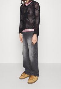 Schwarzer, gestrickter Pullover mit langen Ärmeln und Sternenmuster über einem rot-weiß gestreiften Hemd, kombiniert mit grauen Cargo-Hosen und taupefarbenen Sneakers.