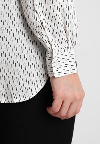 Chemise blanche avec un motif de rayures verticales noires, présentant des manches froncées et des poignets boutonnés. Tissu texturé avec une finition lisse.