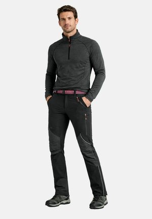 Mann trägt dunkelgrauen Pullover mit Reißverschluss, schwarze Outdoor-Hose mit Reißverschlusstaschen und Wanderschuhe, steht mit Händen in den Taschen.