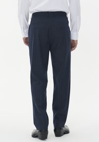 Man som har på sig marinblå randiga byxor, vit skjorta inknuten och svarta dressskor, sedd bakifrån mot en vit bakgrund.