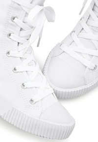 Zapatillas de lona blancas con punta redonda, suela de goma y cordones planos. Presentan ojales metálicos y costuras texturizadas en los lados.