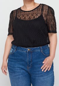Top en dentelle noire à manches courtes, avec un design superposé sur une couche de base unie. Associe à un jean en denim bleu taille haute.