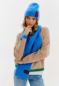 Jasnoniebieska dziana czapka z prążkami z naszywką, dopasowany szalik oraz taupe sweter w zielone i czerwone paski na dolnym brzegu, noszone na dżinsach.