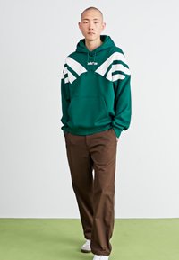 Sweatshirt verde com capuz, com riscas diagonais brancas e bolso frontal, combinado com calças castanhas e ténis brancos num fundo simples.