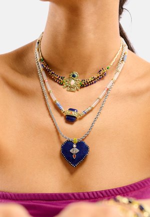 Colliers superposés avec des perles colorées, un pendentif oiseau doré, et un charm en forme de cœur bleu avec des détails complexes et des accents métalliques.