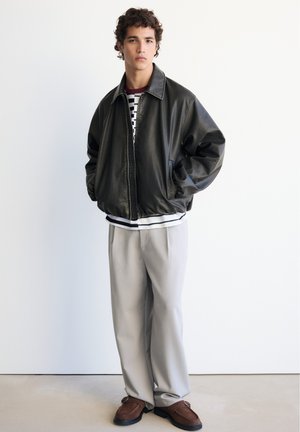Jeune homme aux cheveux bouclés portant une veste en cuir noire, une chemise rayée, un pantalon large beige et des chaussures marron, debout contre un mur blanc.