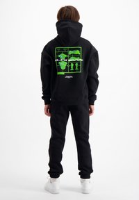 Zwarte hoodie met een groen grafisch ontwerp op de rug, gecombineerd met zwarte sweatpants. Met capuchon, van katoen, snug fit. Witte sneakers.