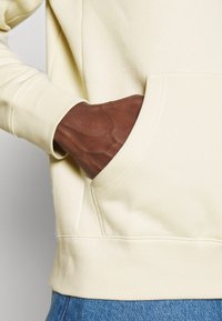 Mörkhyad hand i framfickan på en krämfärgad huvtröja som bärs med blå denimjeans.