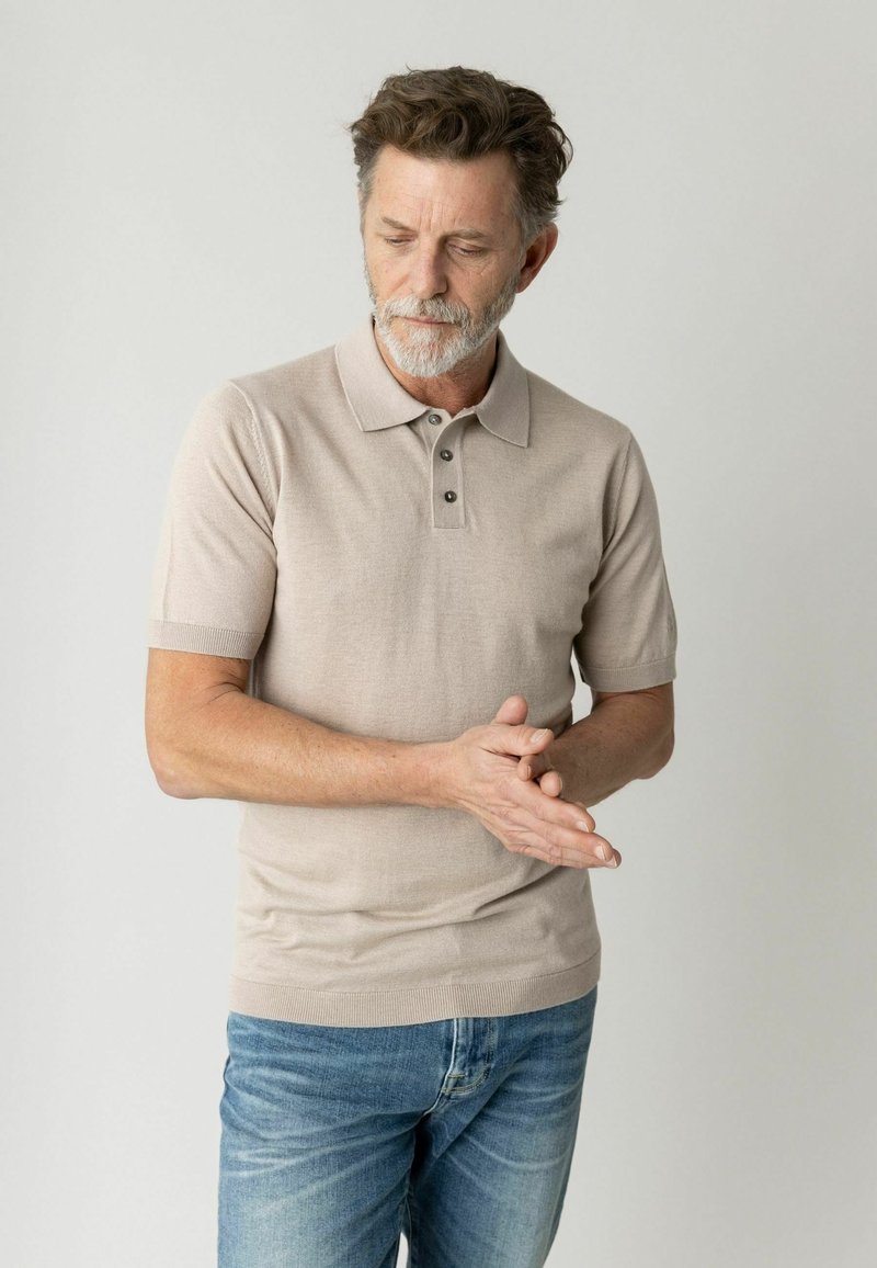 Uomo di mezza età con barba grigia, indossa una polo beige e jeans blu, guarda verso il basso mentre stringe le mani davanti a uno sfondo semplice.