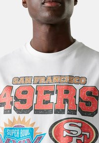 Valkoinen puuvillainen T-paita, jossa on suuria graafisia kuvastoja: "SAN FRANCISCO 49ERS" punaisena ja oranssina, sekä "SUPER BOWL" ja joukkueen logo-koristeet.