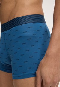 Siniset boxerit pehmeästä kankaasta, joissa on musta raidallinen vyötärö ja toistuva "JOOP!"-logokuosi.