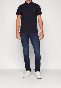 Marinblå polo med krage och plaggknäppning med tre knappar, ihop med mörkblå jeans och vita sneakers, vilket ger en avslappnad look.