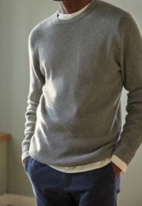 Grauer, gestreifter Pullover mit rundem Ausschnitt, getragen über einem weißen Hemd mit Kragen, kombiniert mit marineblauen, strukturierten Hosen. Weicher, gestrickter Stoff.