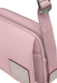 Piccola borsa a tracolla rosa con zip argentata e tracolla abbinata, con un patch "STUDIO BERLIN" sul davanti.