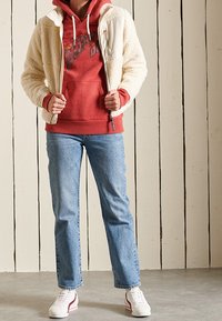Chaqueta de forro polar crema con un panel blanco, sudadera roja con capucha, jeans azules y zapatillas blancas con acentos rojos. Materiales texturizados y un ajuste casual.