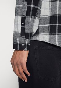 Černá a šedá kostkovaná košile na knoflíky s měkkou texturou, se zapínáním na knoflíky na manžetách a špičatým límcem, kombinovaná s černými džíny.