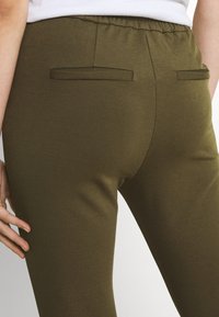 Leggings verde-oliva feitos de tecido elástico, com uma cintura plana e dois bolsos traseiros. Textura suave com design justo.