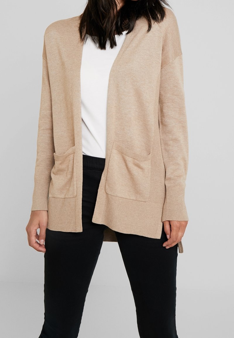 Femme portant un cardigan beige ouvert à l'avant avec des poches, sur une chemise blanche et un pantalon noir, sur un fond uni.