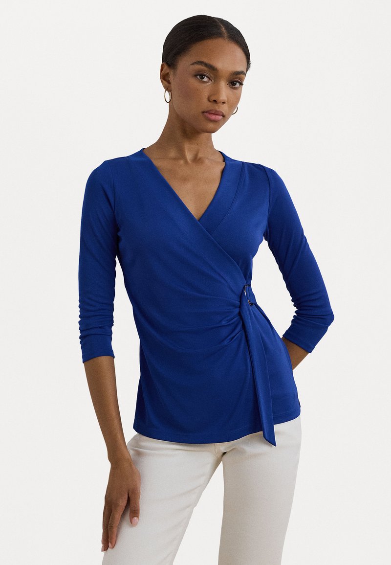 Royal blue, long-sleeve wrap top with a V-neck and gathered side detail. Made from soft fabric, featuring a knot accent at the waist.