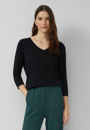 Haut noir à manches longues et col en V, fabriqué en tissu lisse, présentant une coupe décontractée et des manches montées, associé à un pantalon vert.